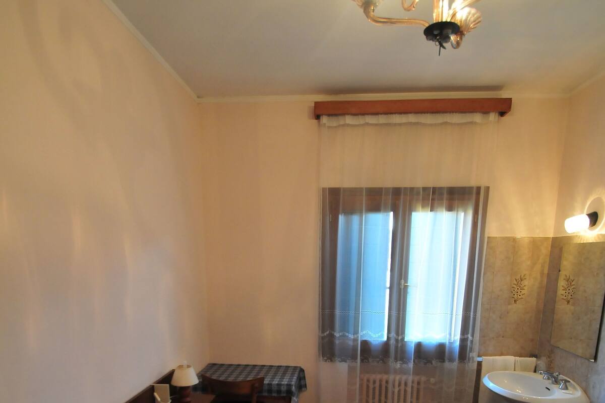 A hotel room with a bed, a table with a lamp, a window with curtains, a sink, and a mirror.