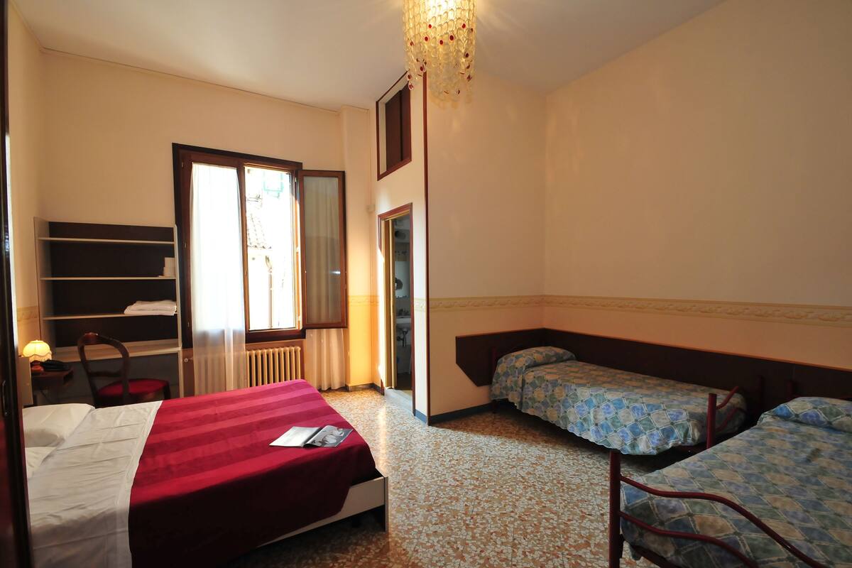 A hotel room with a single bed, two single beds, a chair, a desk with a lamp, a wardrobe, and a ceiling light fixture.