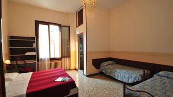 A hotel room with a single bed, two single beds, a chair, a desk with a lamp, a wardrobe, and a ceiling light fixture.