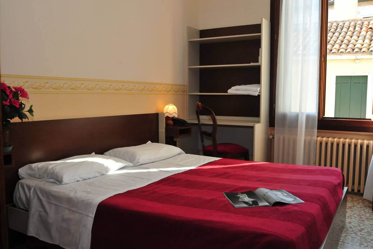 A hotel room with a bed, a nightstand, a chair, a shelf with towels, and a window with a view of the outside.