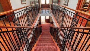 A grand staircase with ornate black railings and a red carpet, leading to a bright room with a large window and a door. - Berg en Zee Guesthouse (Cape Town)