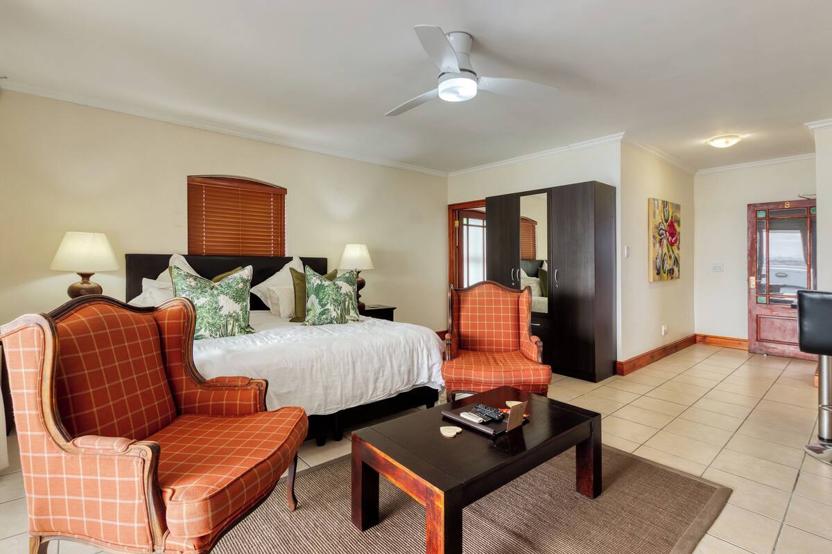 Une chambre d’hôtel comprenant un lit, deux fauteuils, une table basse, un ventilateur de plafond et une porte donnant accès à une salle de bain.