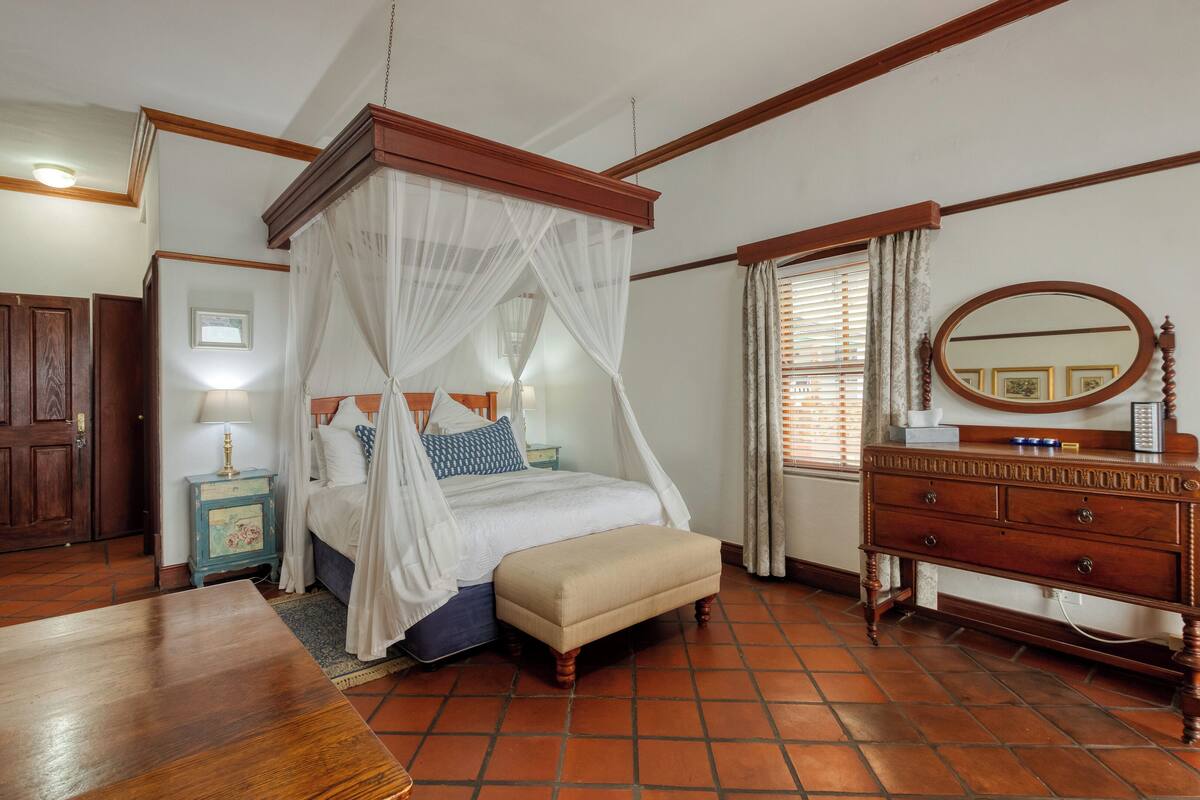 Une chambre à coucher avec un lit à baldaquin, une commode en bois, une table de chevet avec une lampe et une fenêtre avec des rideaux.