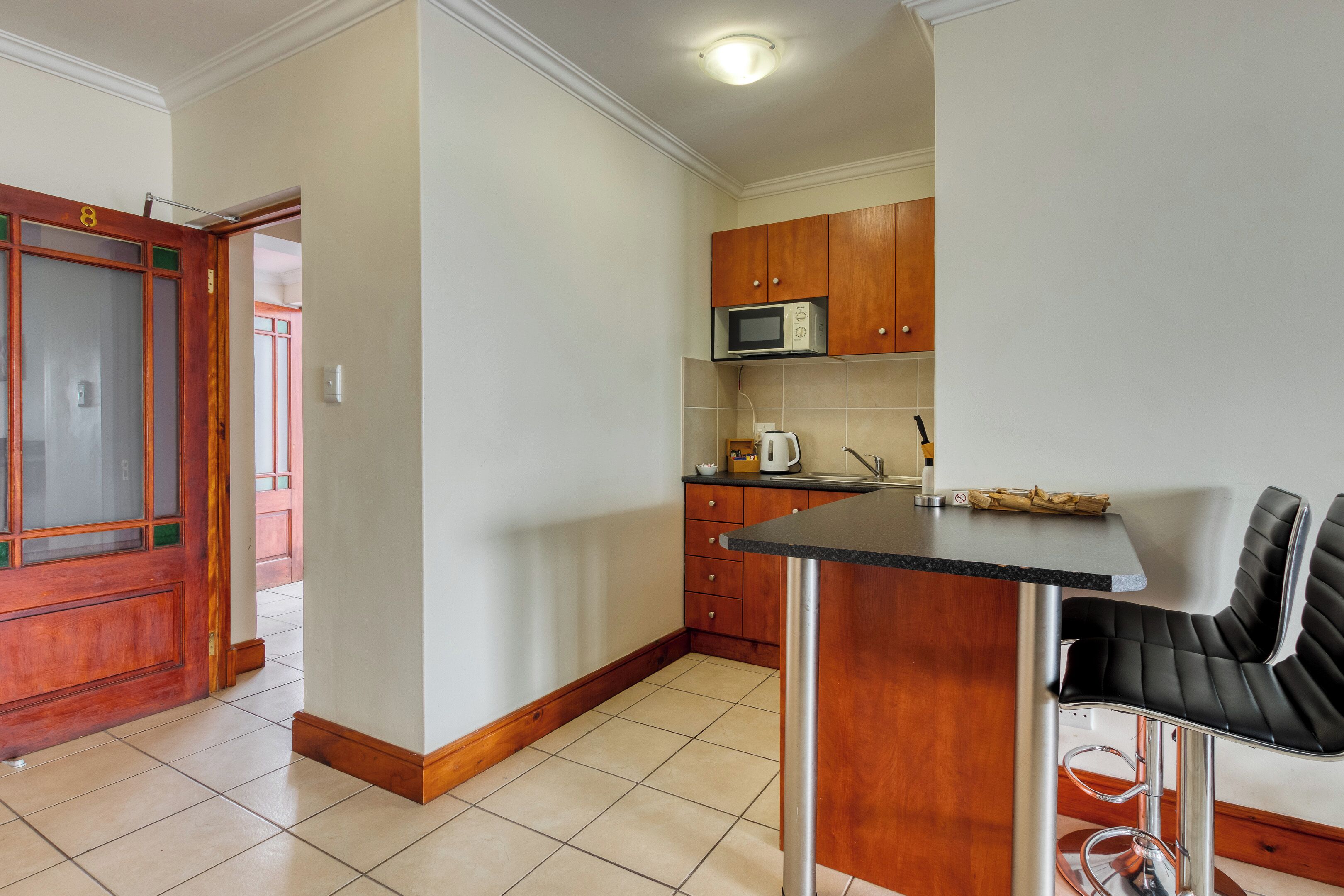 A kitchen with wooden cabinets, a microwave, a kettle, and a bar stool.