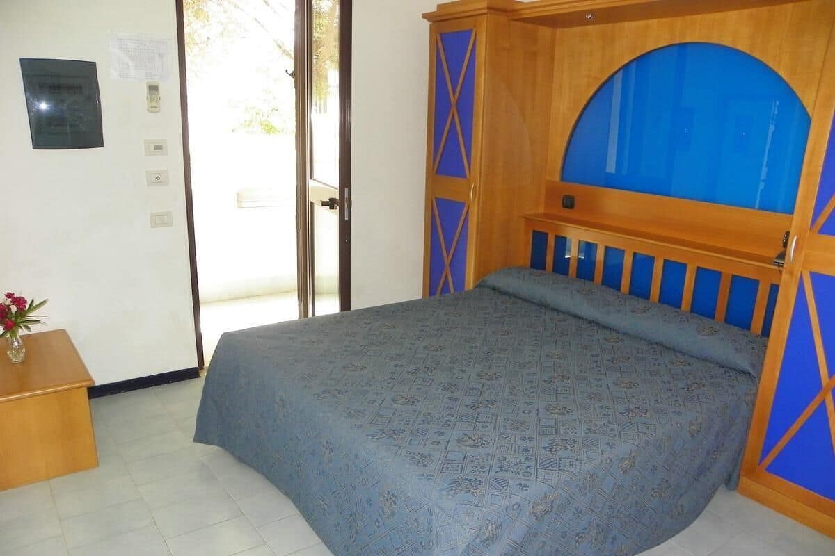 Un dormitorio con cama, cabecera de madera, una mesita de madera con un florero y una puerta que da a un balcón.