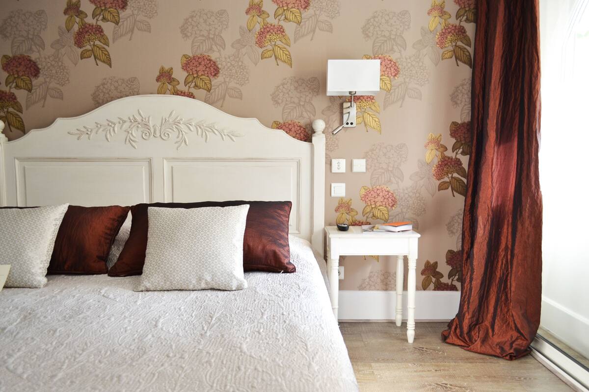 Una cama bien hecha con cabecera blanca y ropa de cama estampada, una mesita de noche pequeña con una lámpara y un diseño de papel tapiz floral.