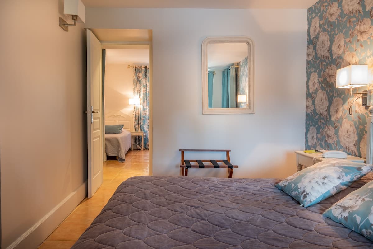 Un dormitorio con una cama grande, una mesita de noche con lámpara, un espejo y una puerta que da a otra habitación.
