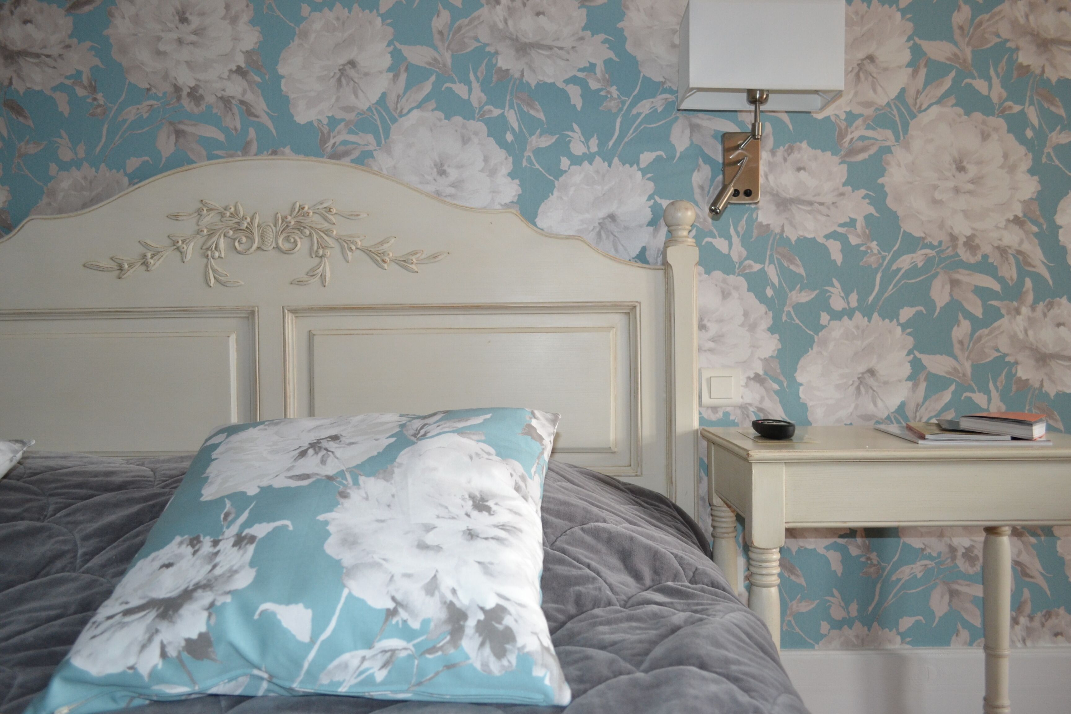 A bedroom with a floral wallpaper, a white headboard, a grey bedspread, and a bedside table with a lamp and books.