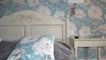 A bedroom with a floral wallpaper, a white headboard, a grey bedspread, and a bedside table with a lamp and books.