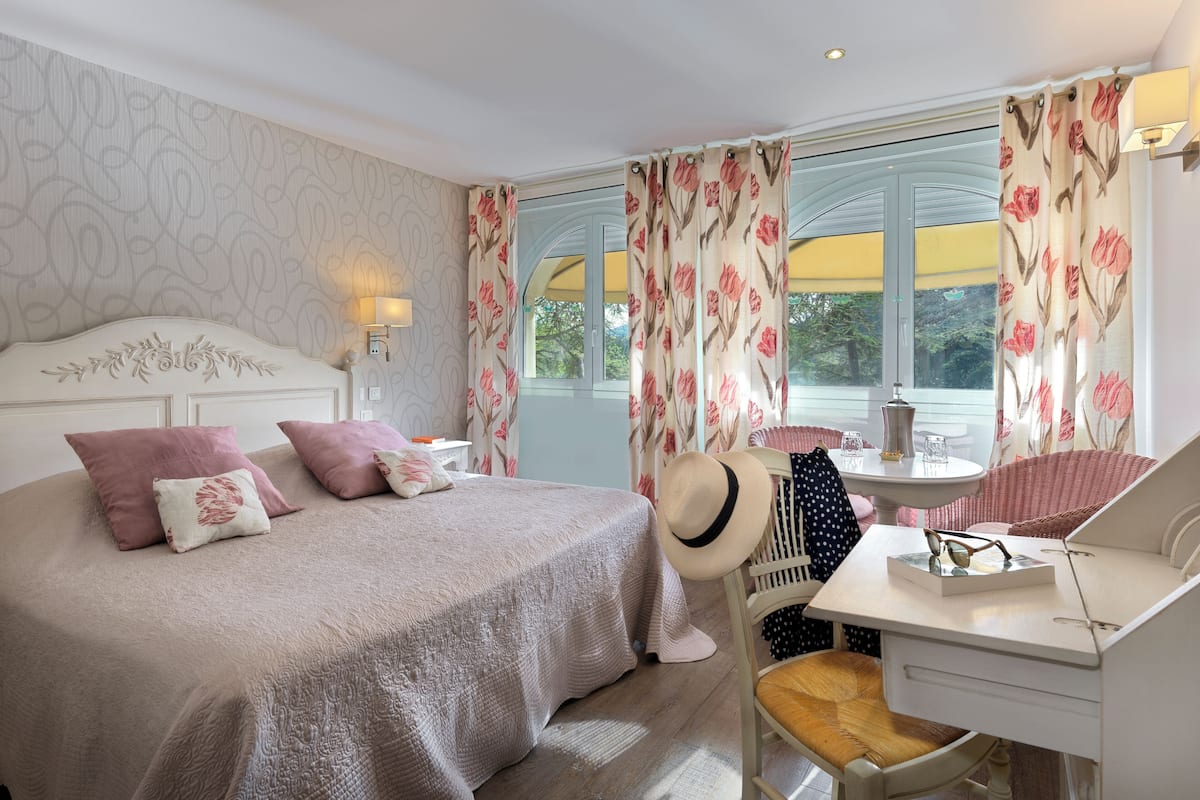 Un dormitorio con una cama grande, un escritorio y una silla. Hay una ventana con cortinas, una lámpara y un sombrero en la silla.