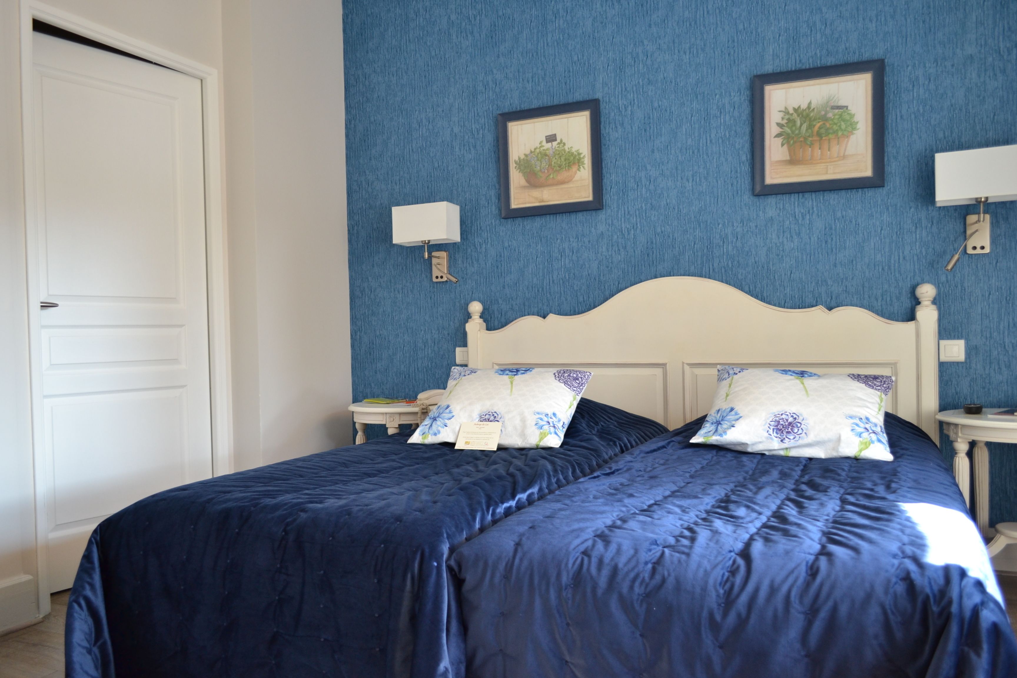 Un lit double avec une tête de lit blanche et une parure de lit bleue, deux tables de chevet avec des lampes et deux tableaux encadrés accrochés au mur.