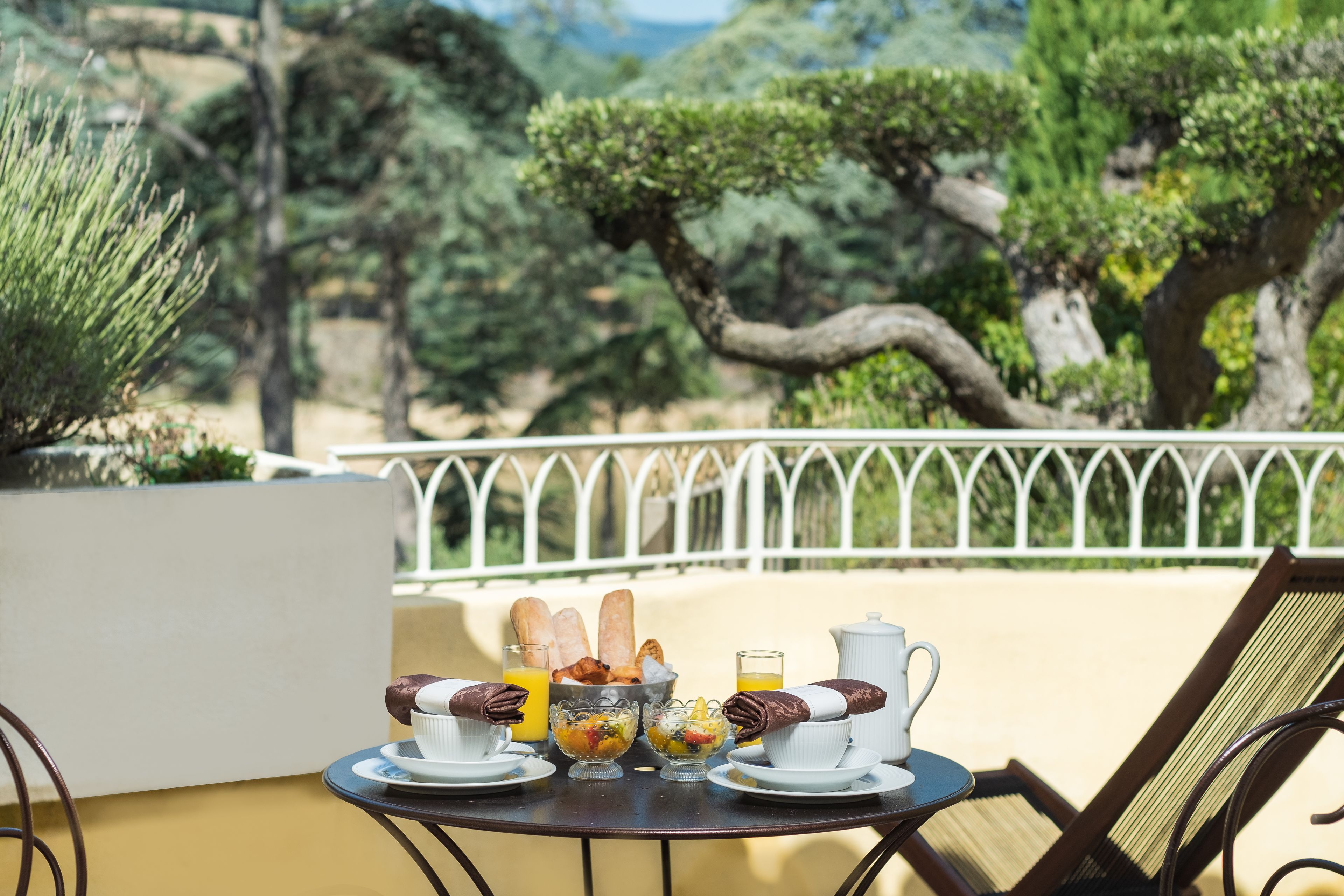 A table set with breakfast items including croissants, fruit, and coffee, overlooking a garden with trees and a white fence.