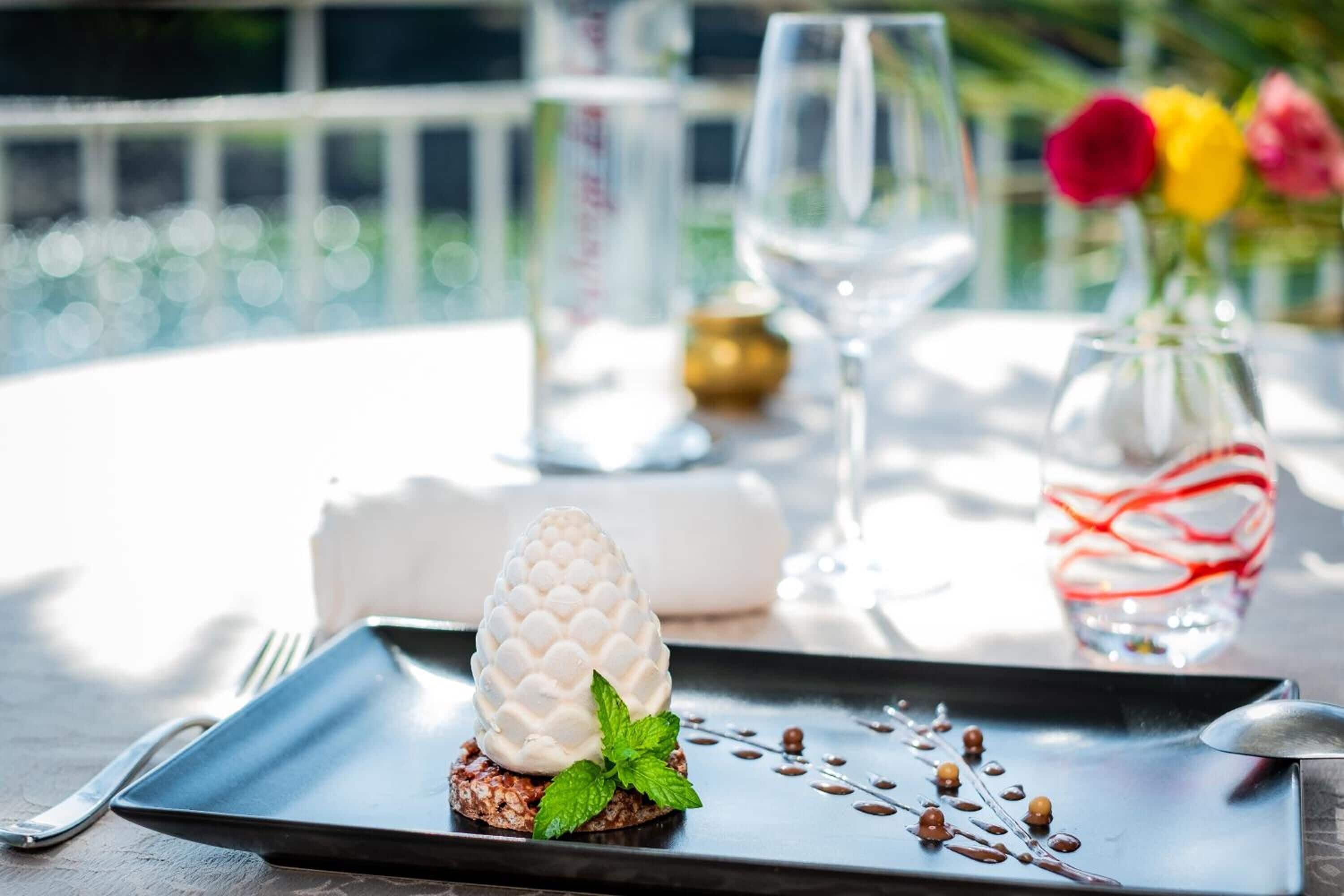 Un dessert servi dans une assiette noire, agrémenté de quelques feuilles de menthe, un verre de vin blanc et un vase rempli de fleurs, le tout posé sur une table.
