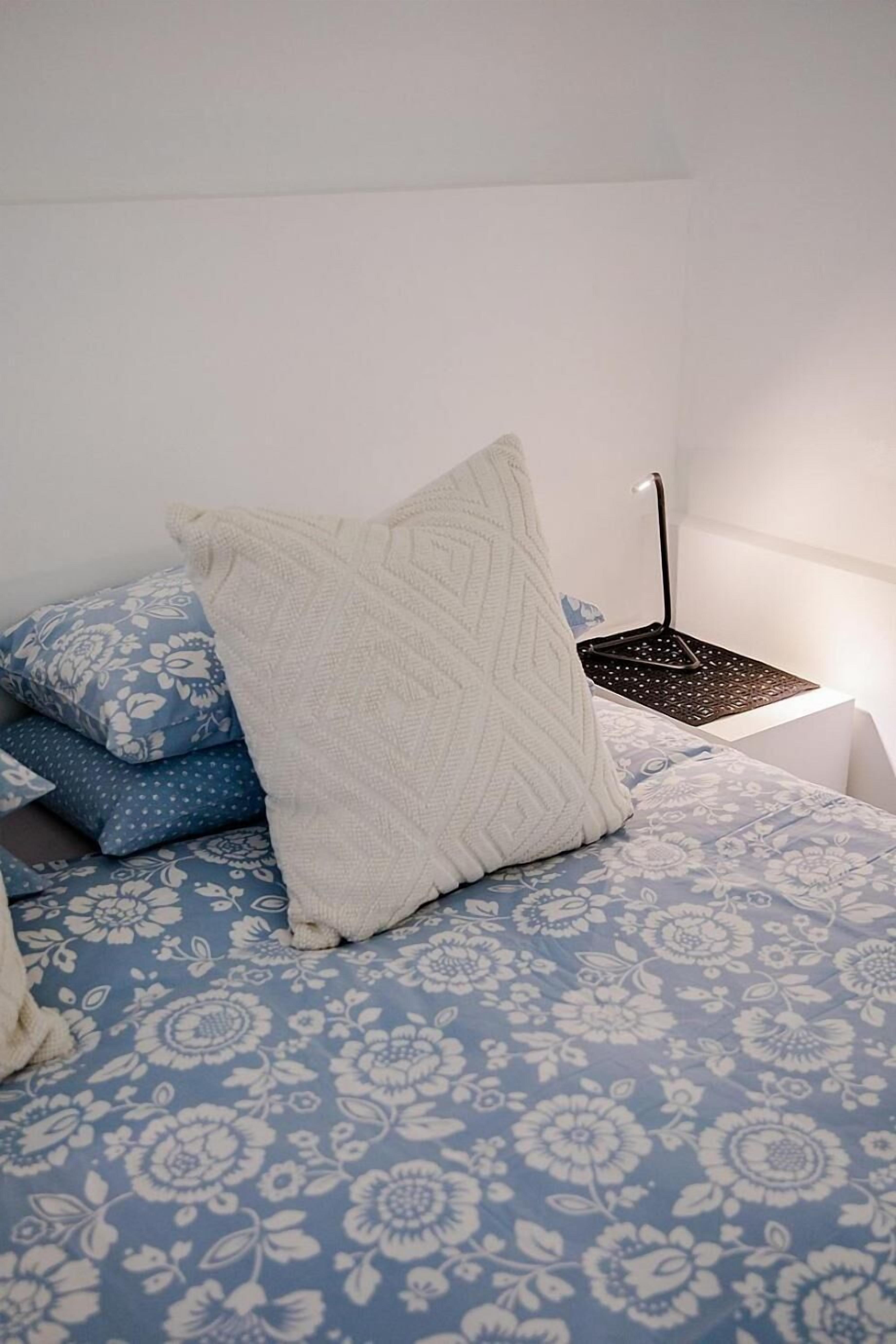 A bed with a blue floral bedspread, a white knitted throw, and a black lamp on a white bedside table.