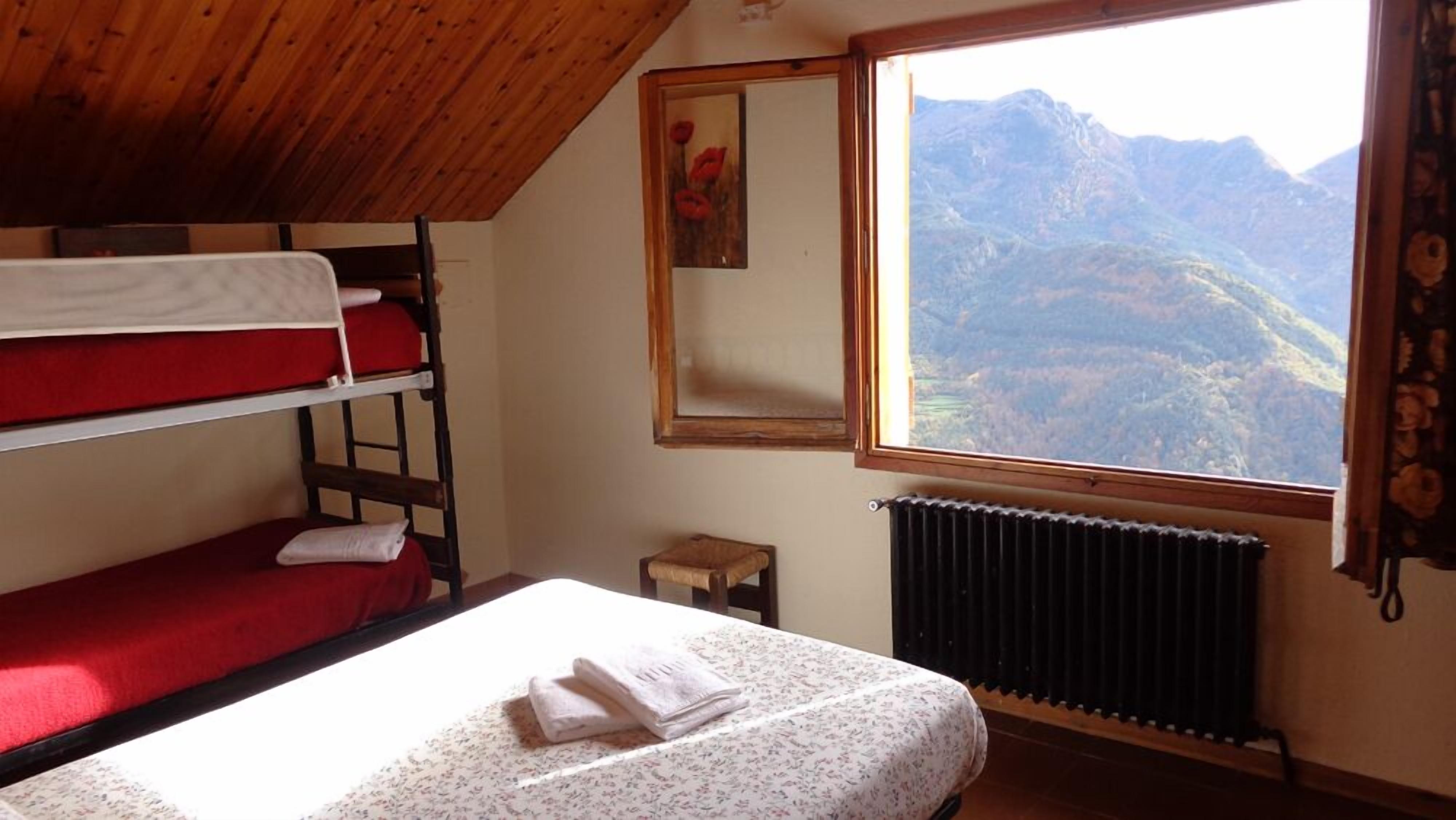 A dormitory room with bunk beds, a small table, a radiator, and a window with a mountain view.