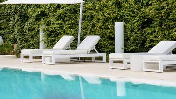 Zona de piscina con sillones blancos y sombrillas, rodeada de abundante follaje.