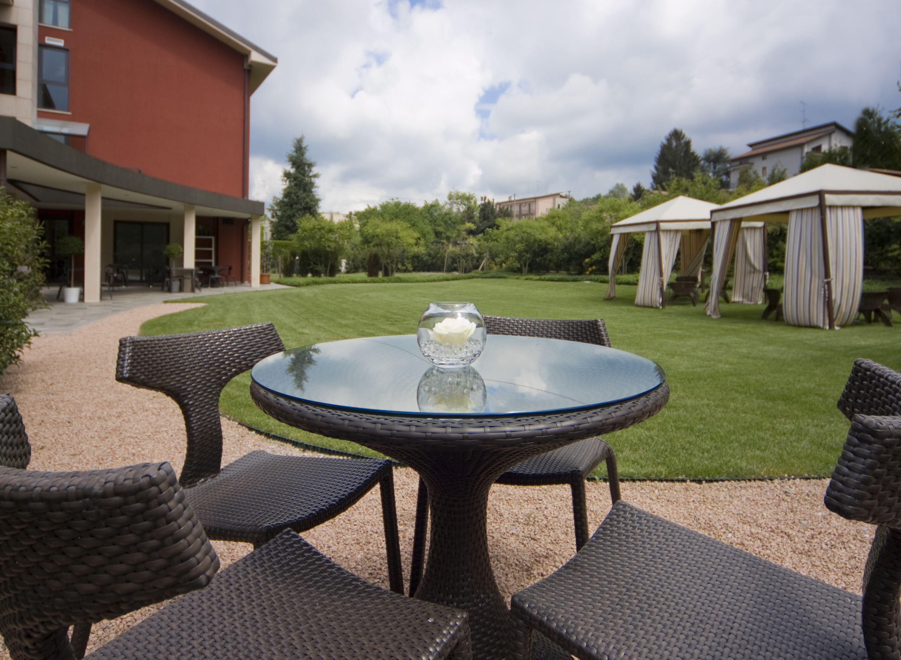 A round glass table with a glass bowl centerpiece, surrounded by wicker chairs, set on a gravel path with a lawn and buildings in the background.