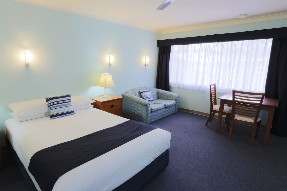 A hotel room with a large bed, two bedside tables with lamps, a chair, a small table, and a window with curtains.