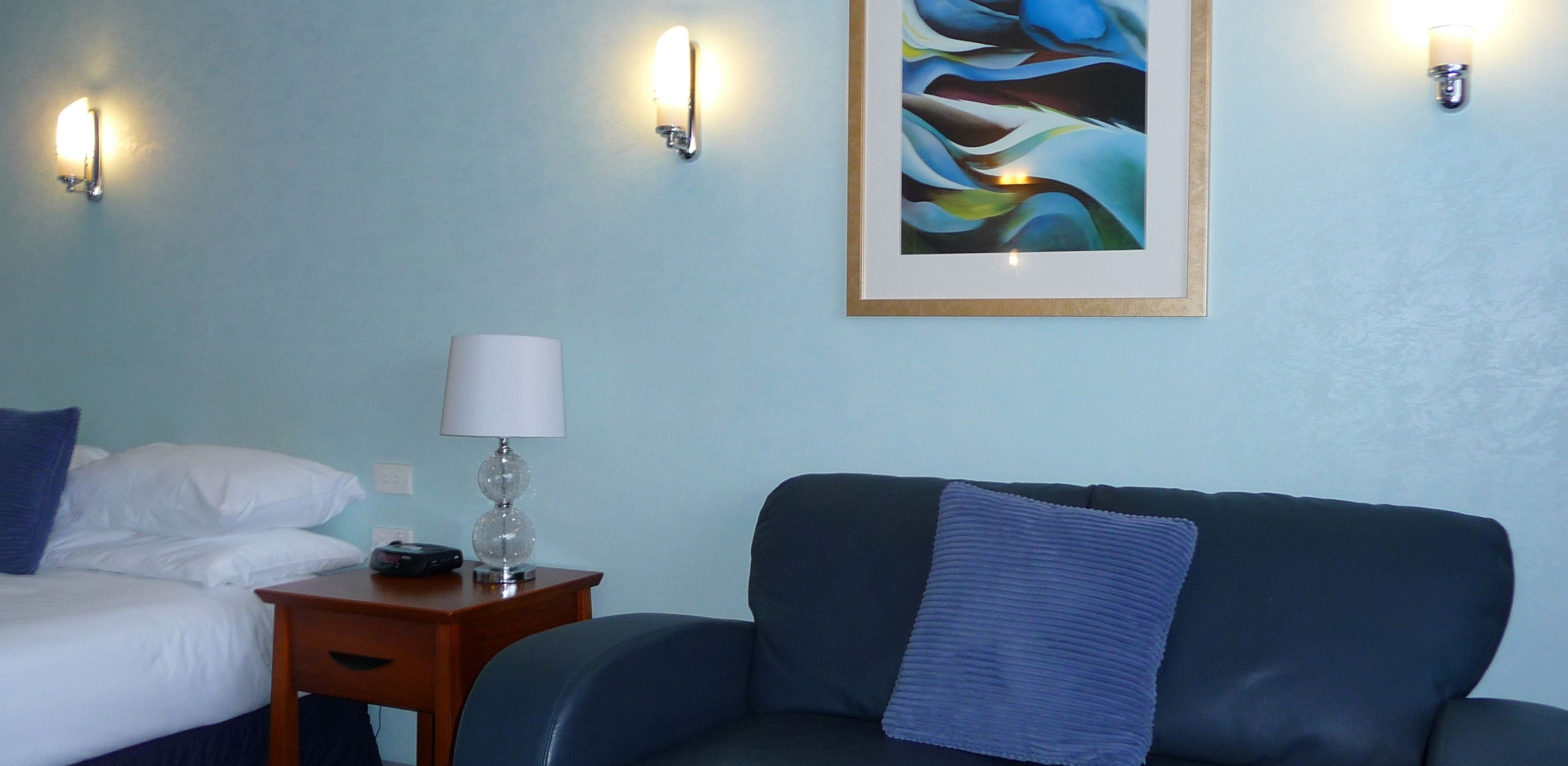 A room with a bed, a small table with a lamp, a couch, and a painting on the wall.