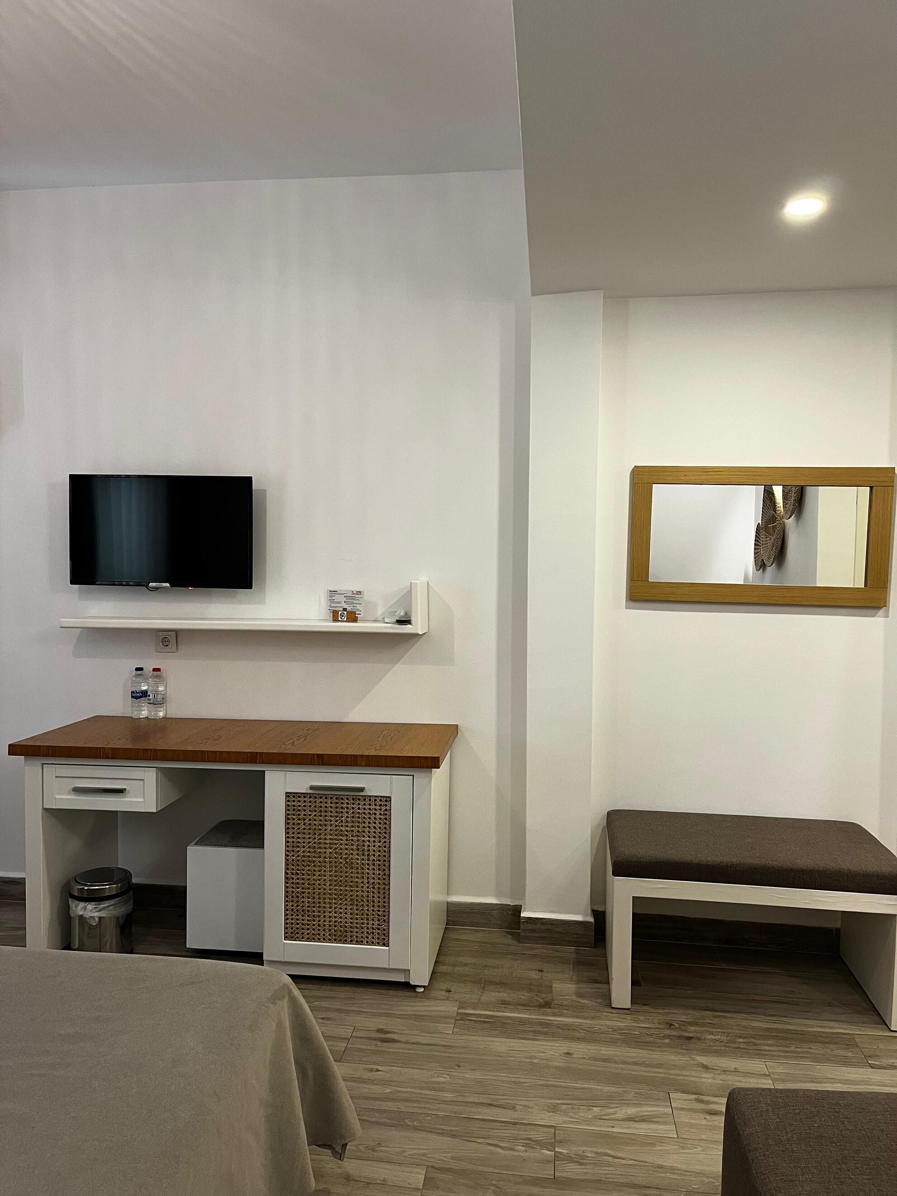 Una habitación de hotel moderna con un escritorio de madera, un televisor de pantalla plana, un estante pequeño con artículos de aseo, un banco y un espejo en la pared.