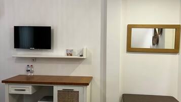 Una habitación de hotel moderna con un escritorio de madera, un televisor de pantalla plana, un estante pequeño con artículos de aseo, un banco y un espejo en la pared.