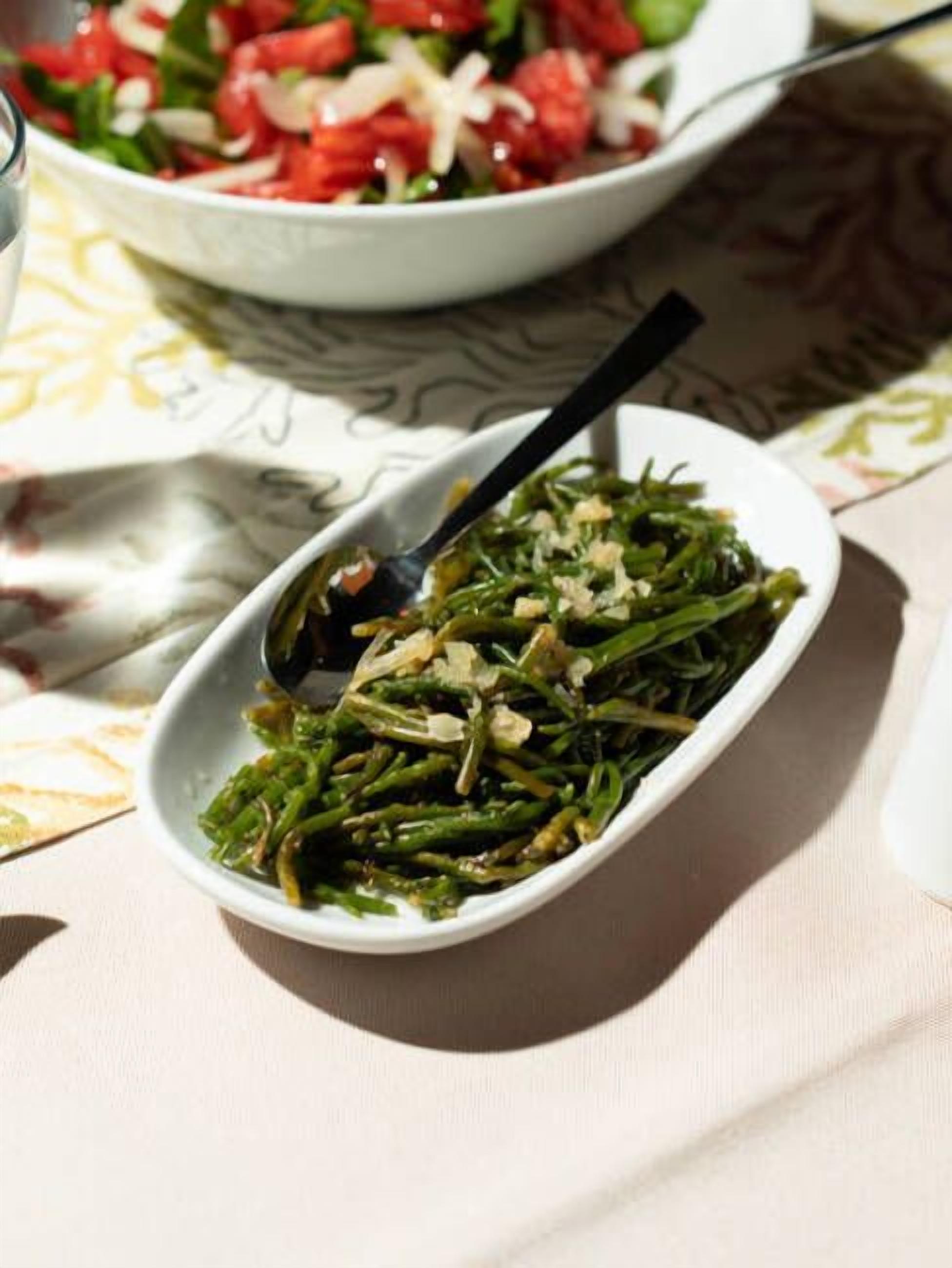 Un tazón blanco con verduras verdes y una cuchara, sobre una mesa con mantel estampado.