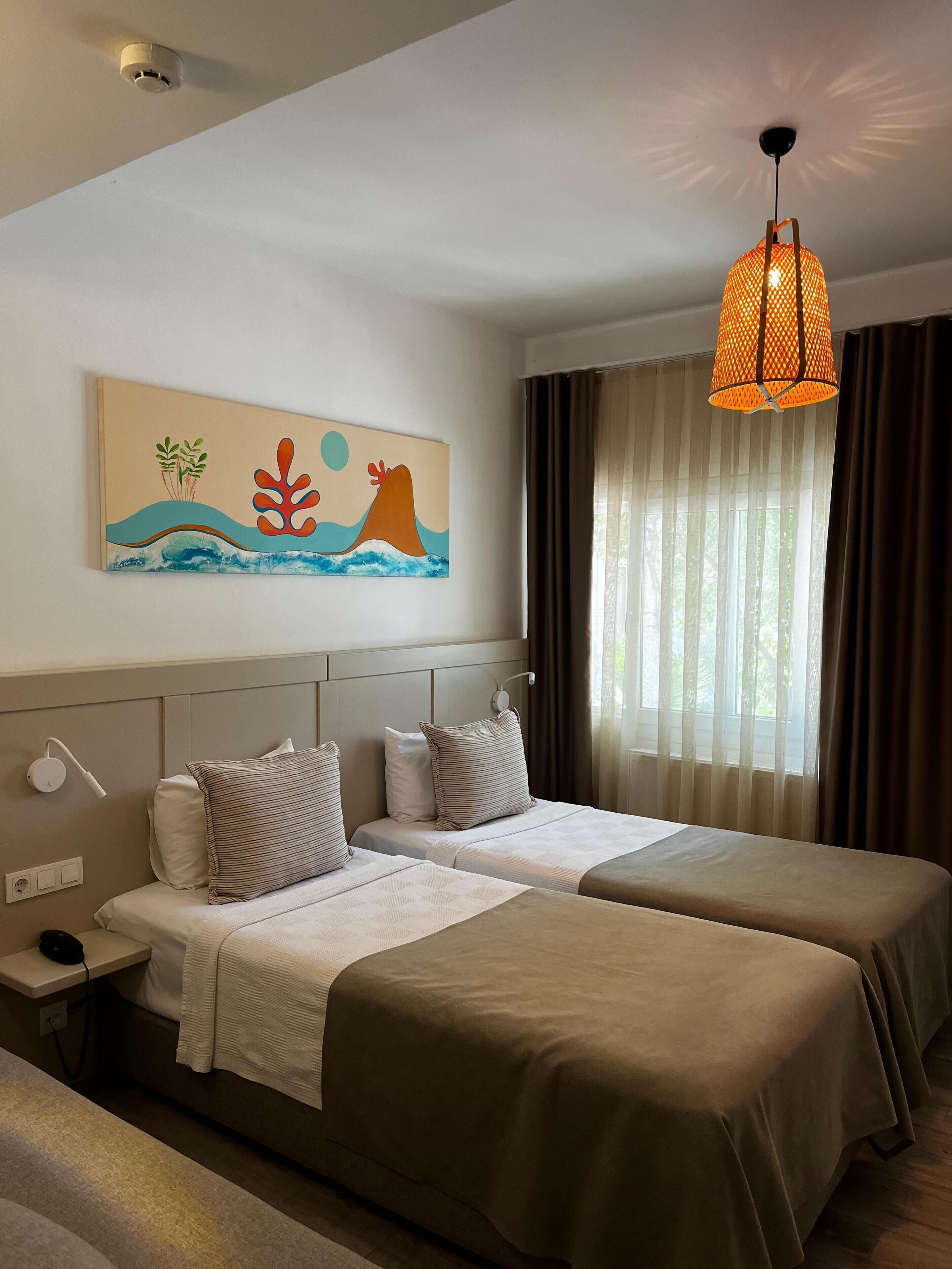 Une chambre d’hôtel avec deux lits, un tableau représentant une scène de plage, un luminaire au plafond et une fenêtre avec des rideaux.