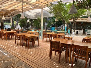 Un espacio de comedor al aire libre con mesas y sillas de madera, una zona de estar techada y lámparas colgantes.