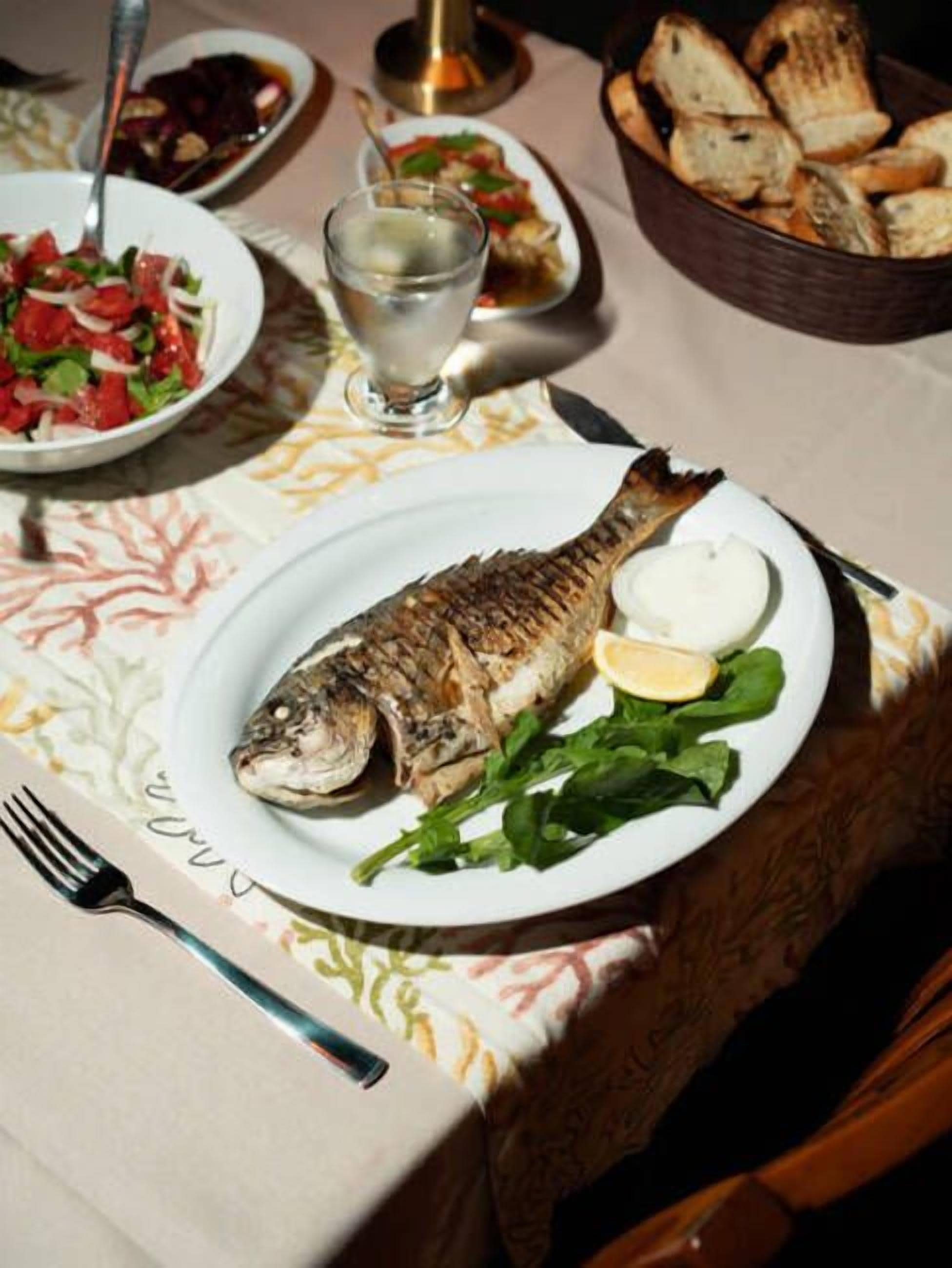 A dining table with a plate of grilled fish, a glass of water, and a basket of bread.