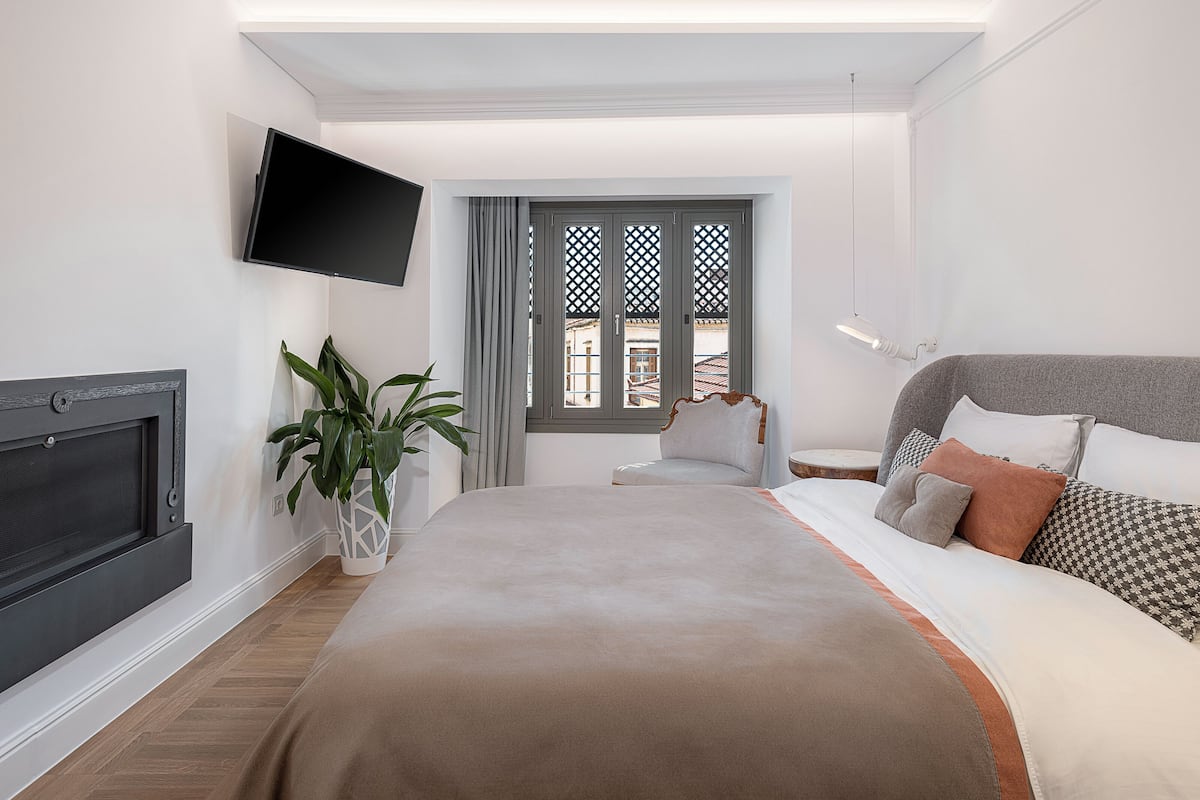 A modern hotel room with a large bed, a TV mounted on the wall, a potted plant, and a window with decorative lattice.