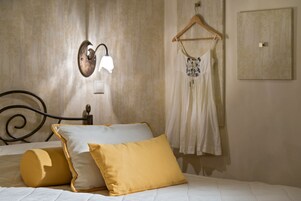 Uma cama bem arrumada com cabeceira, luminária fixada na parede, roupa pendurada e um quadro na parede.