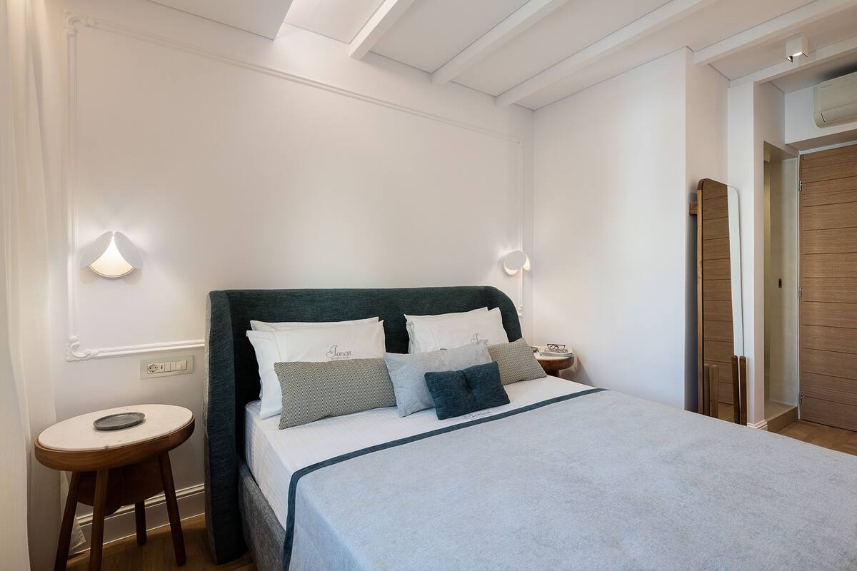 A neatly made bed with a dark headboard and multiple pillows, a bedside table with a lamp, and a wooden door in the background.