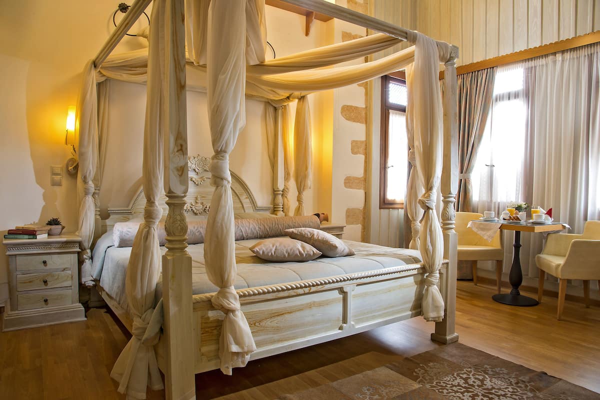 A four-poster canopy bed with a wooden frame and draped curtains, a bedside table with a lamp, and a small table with chairs in the room.