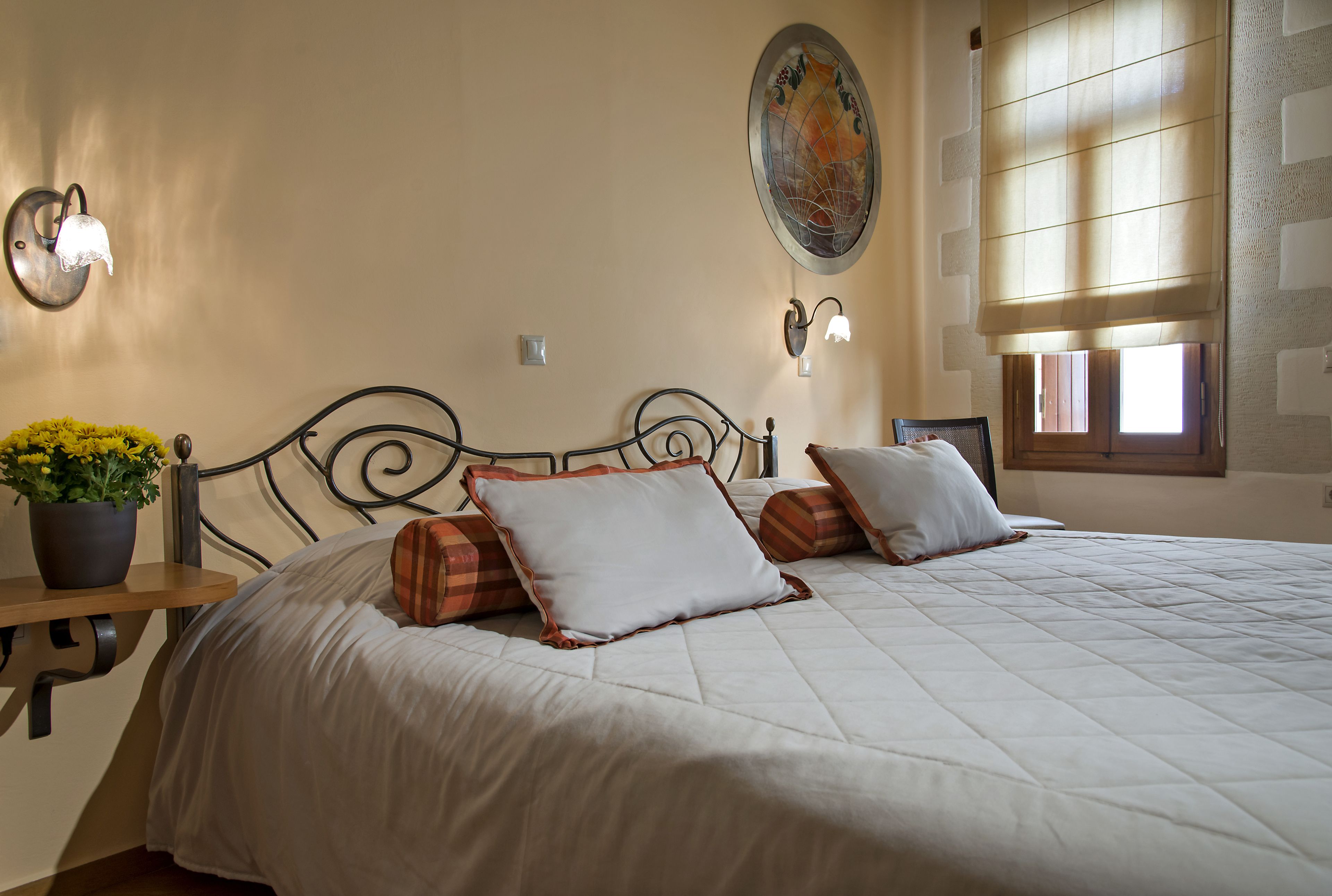 A neatly made bed with white bedding and two pillows, a potted plant on a wooden nightstand, and a wall-mounted light fixture.