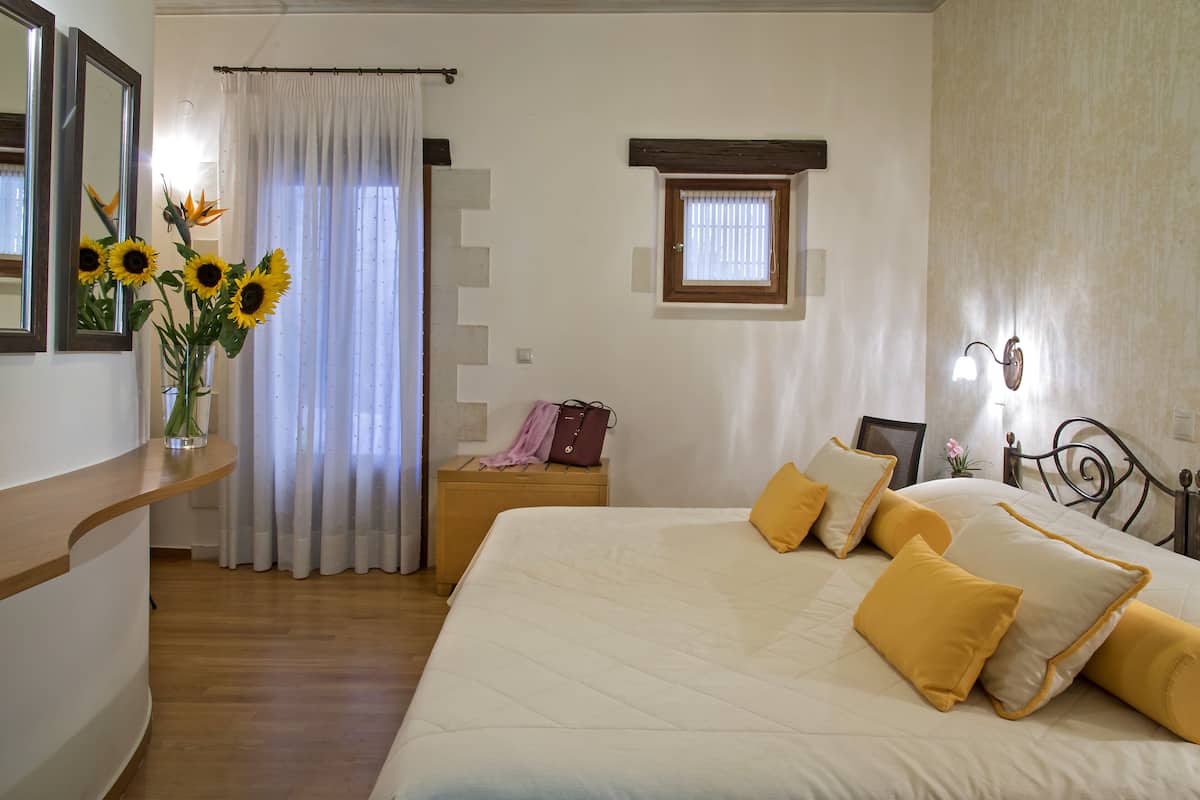 A bedroom with a large bed, a nightstand, a dresser, a window with curtains, and a vase with sunflowers.
