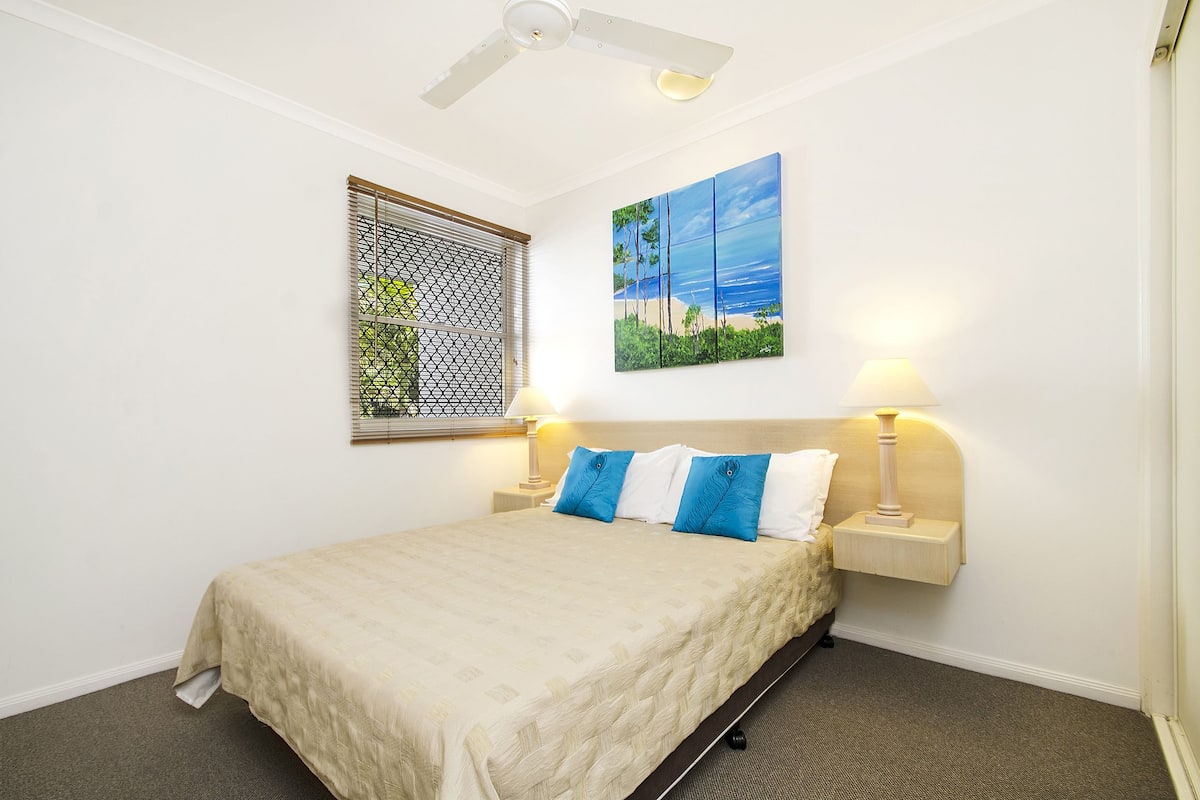 A bedroom with a bed, a ceiling fan, a window with a metal screen, and a wall-mounted painting.