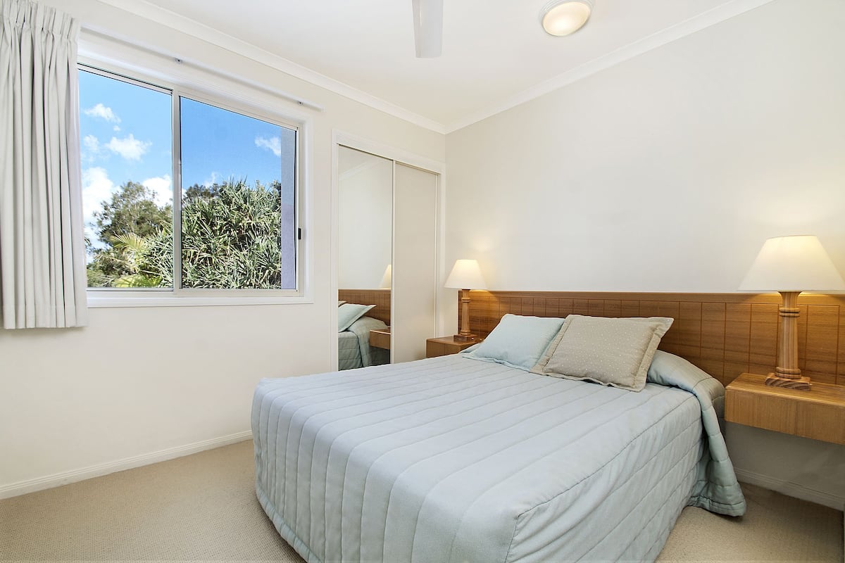 A bedroom with a large bed, two bedside tables with lamps, a window with curtains, and a view of trees outside.