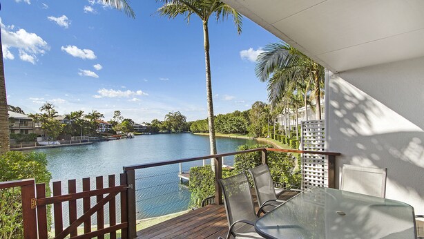 A balcony with a glass table and chairs overlooking a lake, surrounded by palm trees and a white fence. - Portside Noosa Waters (Noosaville)