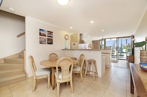 A dining area with a round table and six wicker chairs, a kitchen with a refrigerator and stove, and a staircase leading to another level. - Portside Noosa Waters (Noosaville)
