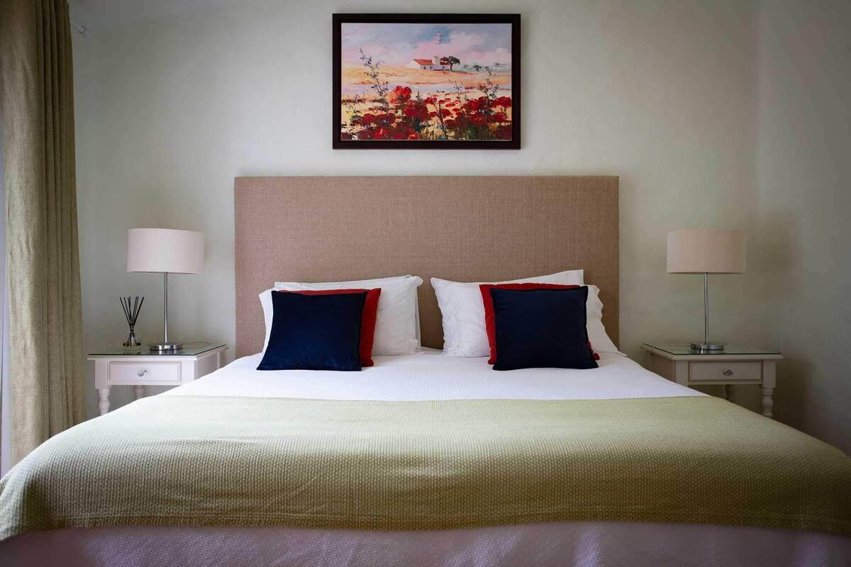 Una habitación de hotel con una cama grande, dos mesitas de noche, un cuadro en la pared y una ventana con cortinas.