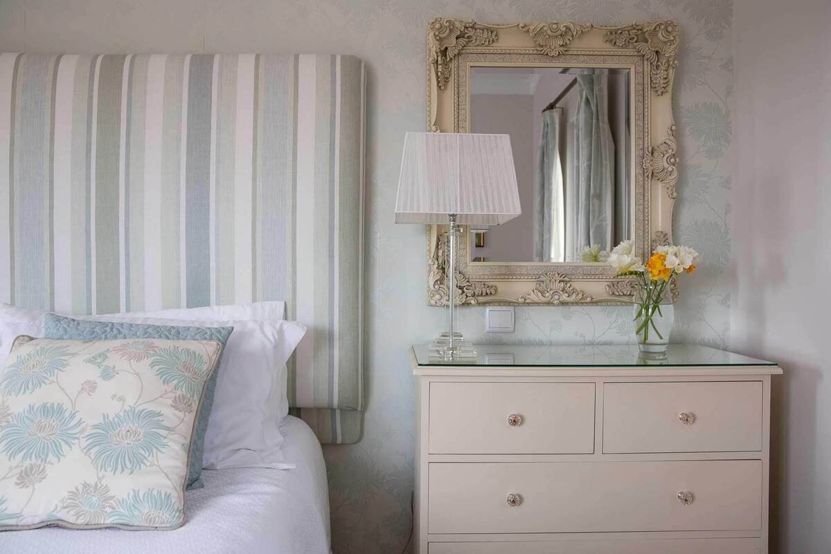 Un dormitorio con cama, mesita de noche, espejo y un florero con flores.