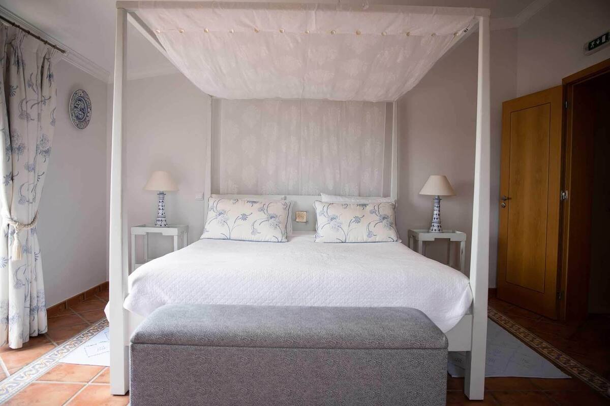 Una cama con dosel y cuatro columnas, con almohadas a juego, dos lámparas de noche y un banco al pie de la cama.