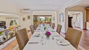 A dining area with a long table set for a meal, surrounded by wicker chairs, with a view of the outdoors through large windows. - Quinta Bonita Luxury Boutique Hotel (Lagos)