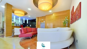 Un hall de hotel con una recepción, un sofá rojo y blanco, un sofá blanco grande, una obra de arte colgada en la pared y un cartel con el nombre del hotel.