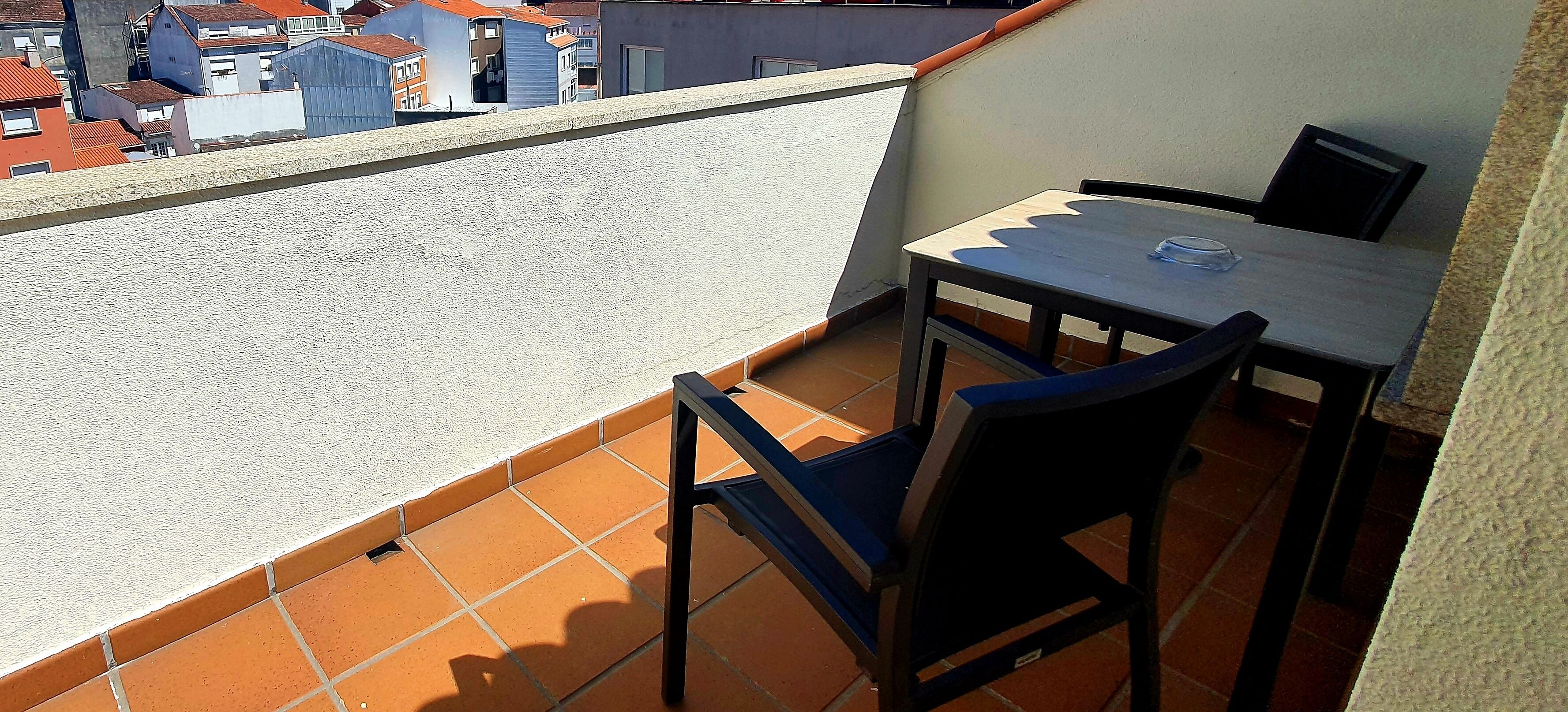 A small balcony with a table and chairs, a tiled floor, and a view of buildings in the background.
