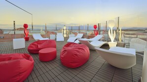 Una terraza en la azotea con sillones reclinables blancos y rojos, pufs y barandilla de vidrio con vistas a un paisaje urbano.