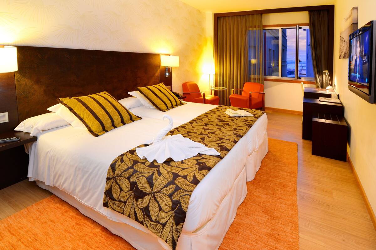 A hotel room with a large bed, two bedside tables, a desk with a chair, a TV, and a window with curtains.