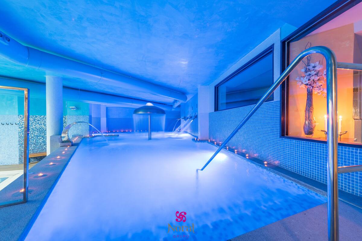 An indoor swimming pool with a blue-lit ceiling, a glass wall with a decorative vase, and a tiled floor.