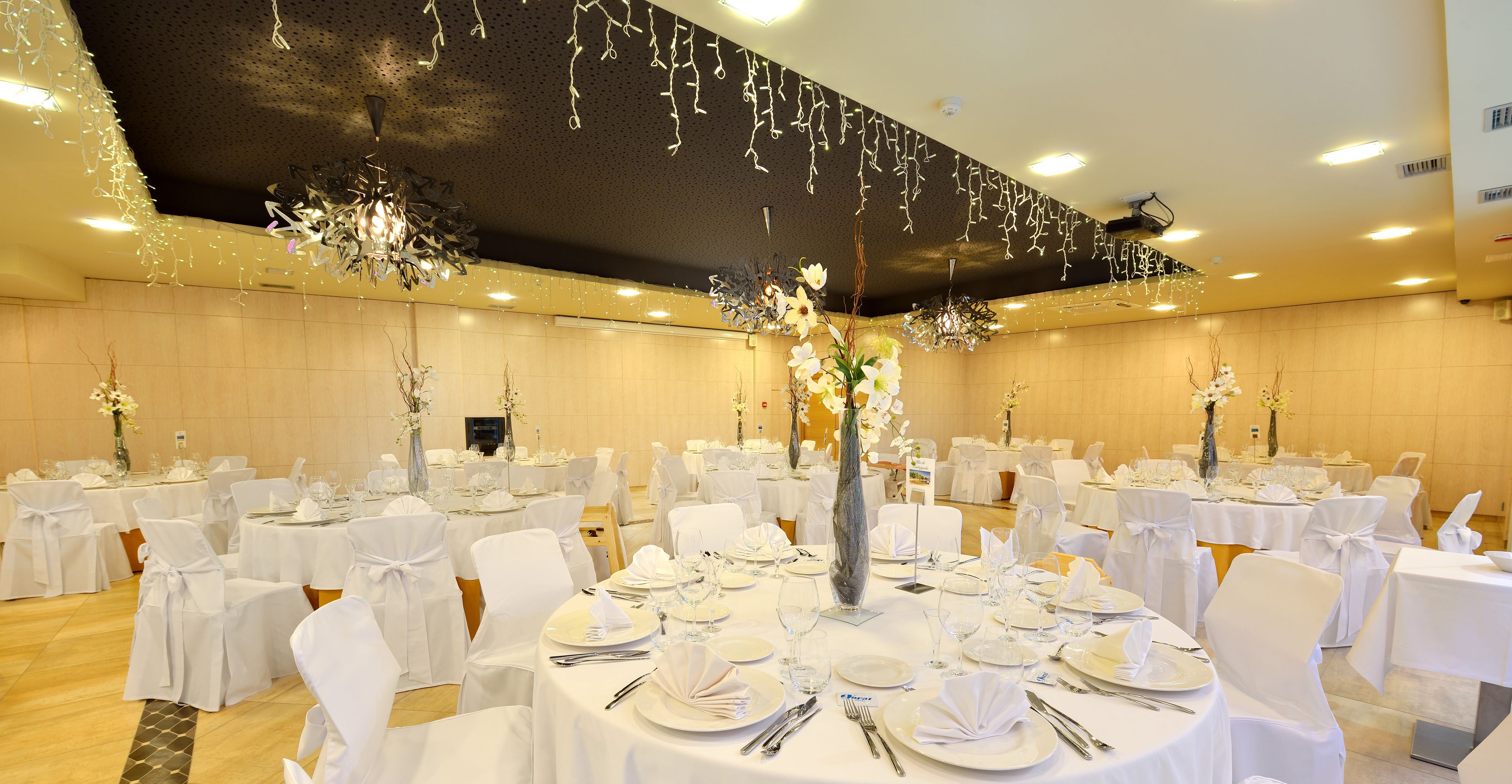 Un salón de banquetes con mesas redondas dispuestas para cenar, sillas blancas y centros de mesa florales.