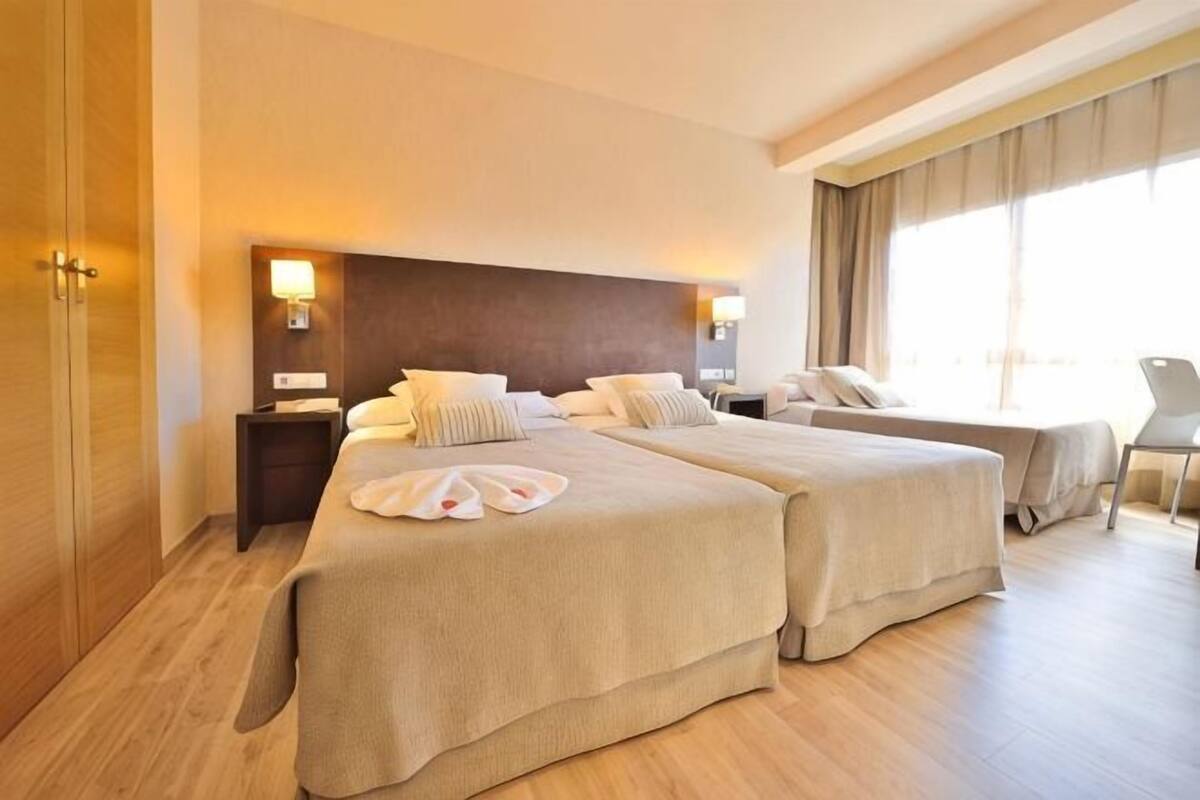 A hotel room with two beds, a wooden floor, a large window, and a chair.